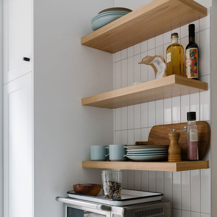 Light oak floating shelves with plates, mugs, glass jar, cutting board, and bottles against white tiled kitchen wall.