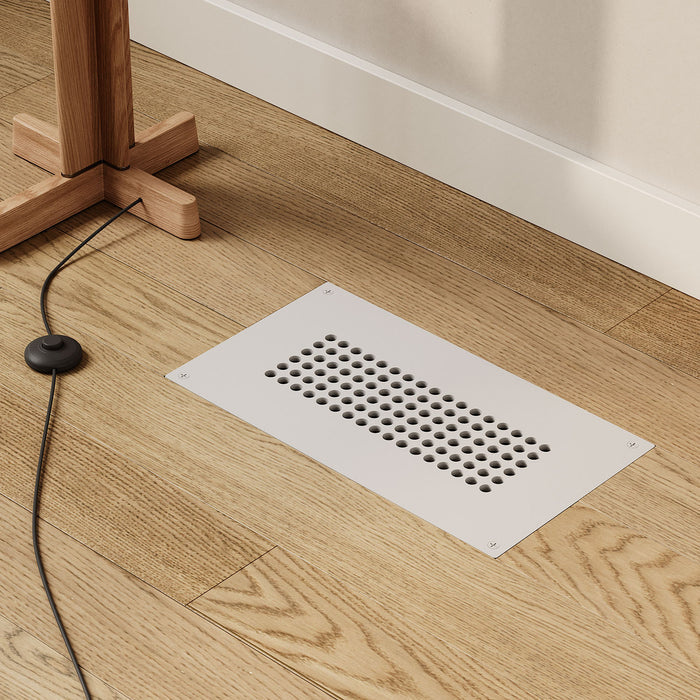 Fritz Vent Cover in matte white steel with a modern perforated pattern, flush-mounted on natural wood flooring near a wooden furniture leg and black cable.