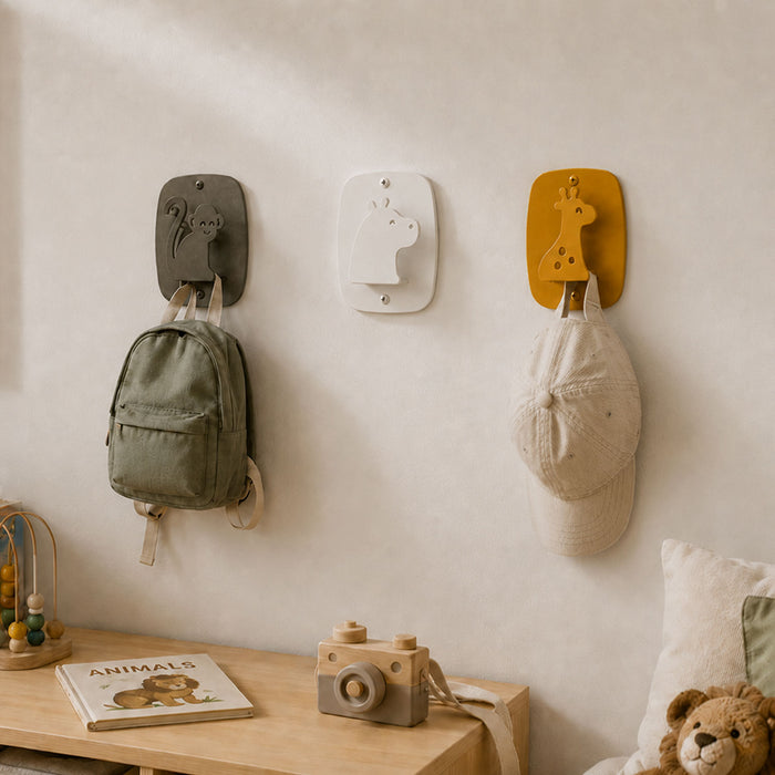Three Kiddo Hooks mounted on a wall in a children's room, featuring playful animal designs holding a backpack and a cap above a wooden shelf with toys and a book.