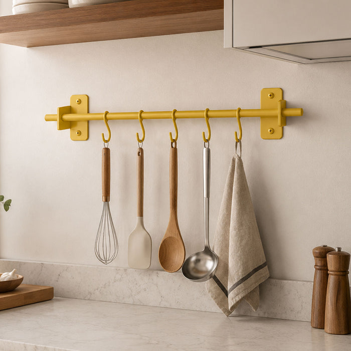 Rue Utensil Rack in matte yellow metal mounted on a kitchen wall holding wooden and stainless steel utensils and a hanging towel.
