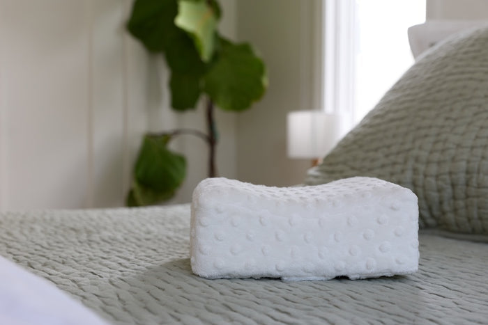textured cream bowroo nursing pillow on a bed with a plant and lamp in the background
