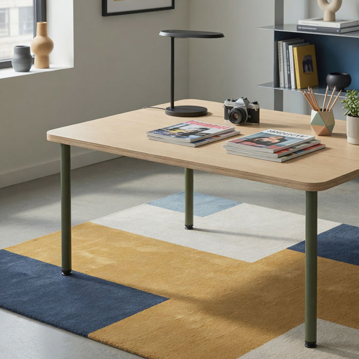 Uppity Table Leg in olive green steel supporting a light wood tabletop with rounded edges, styled with magazines, a camera, and a black modern table lamp on a geometric patterned rug.